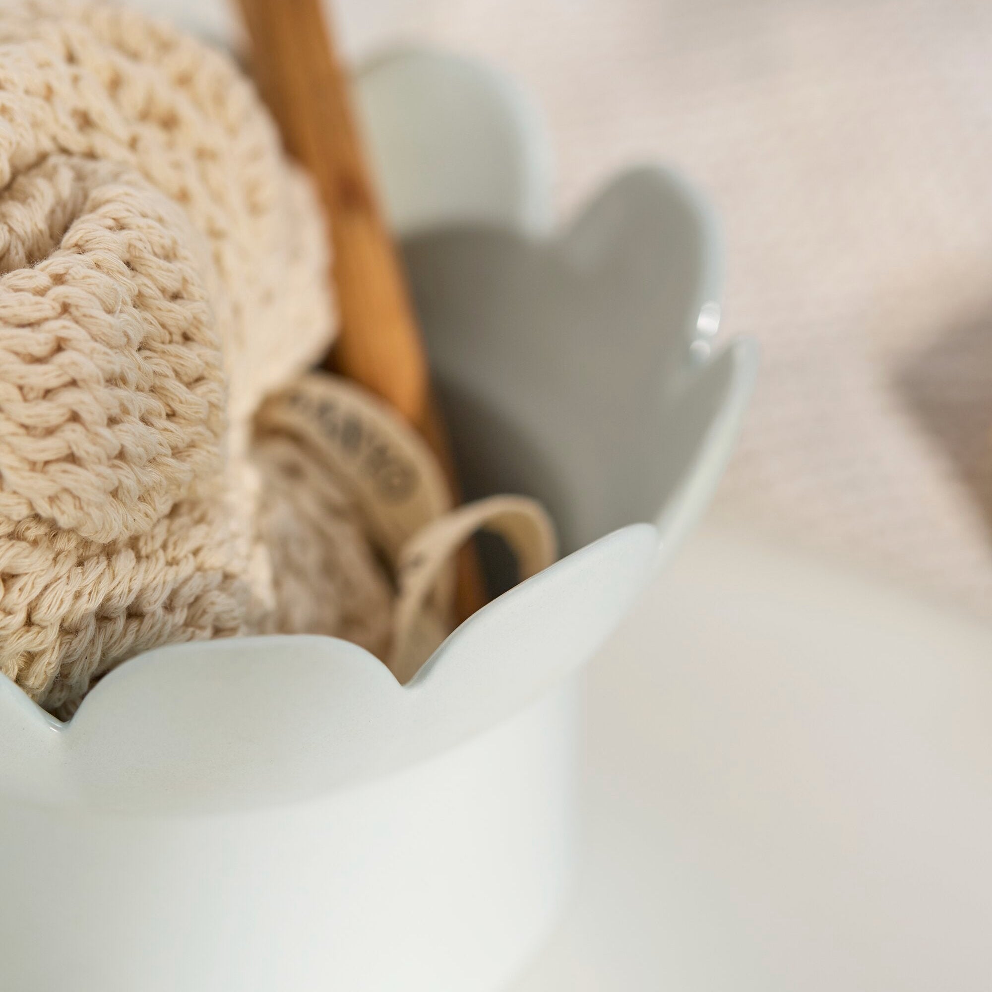 A close-up of a white ceramic toothbrush holder with scalloped edges, containing a beige knit washcloth and a wooden toothbrush, set on a soft white surface.