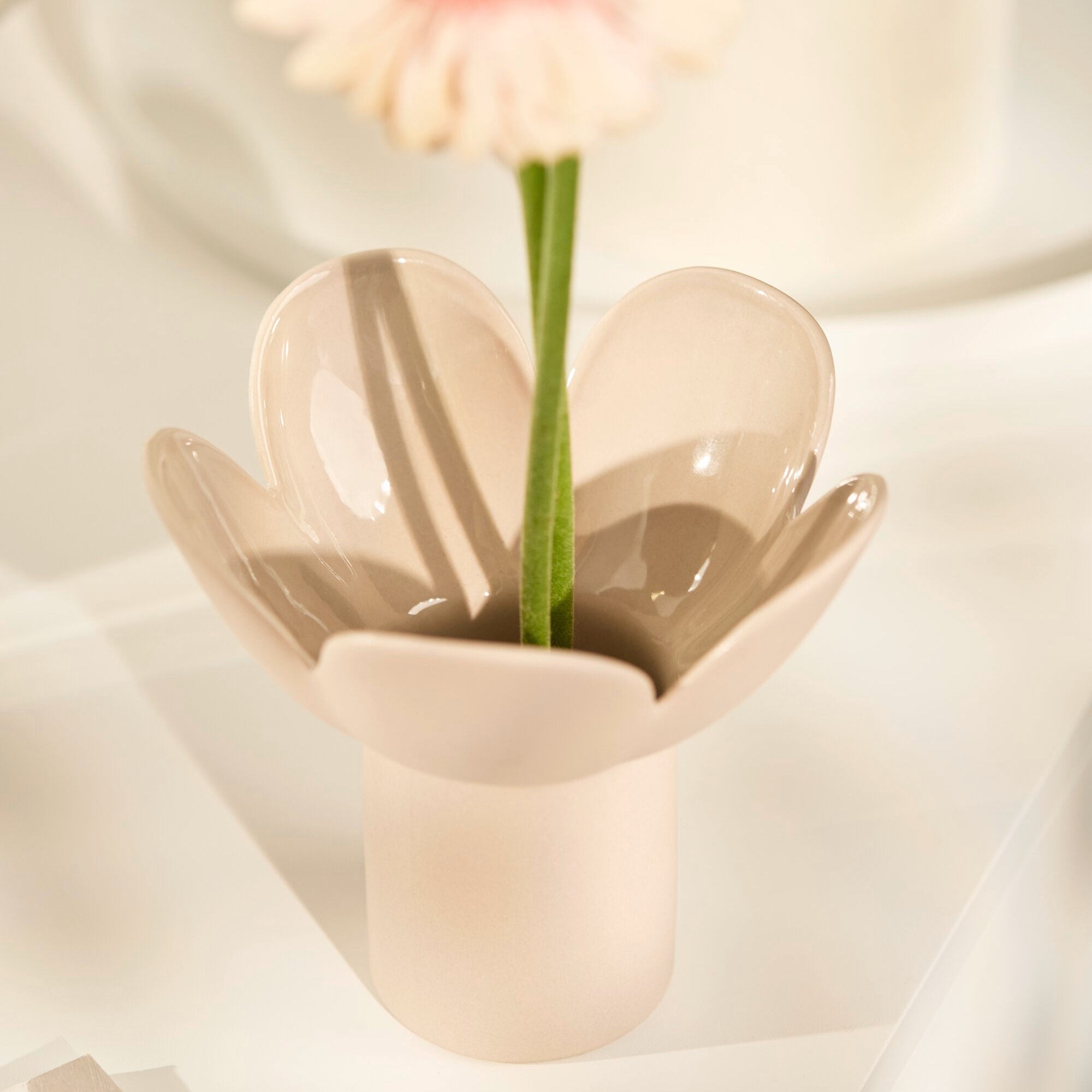 A minimalist ceramic flower-shaped vase in a warm cream-beige color, holding a single pink-and-white daisy with a green stem, set against a soft, draped white fabric background.