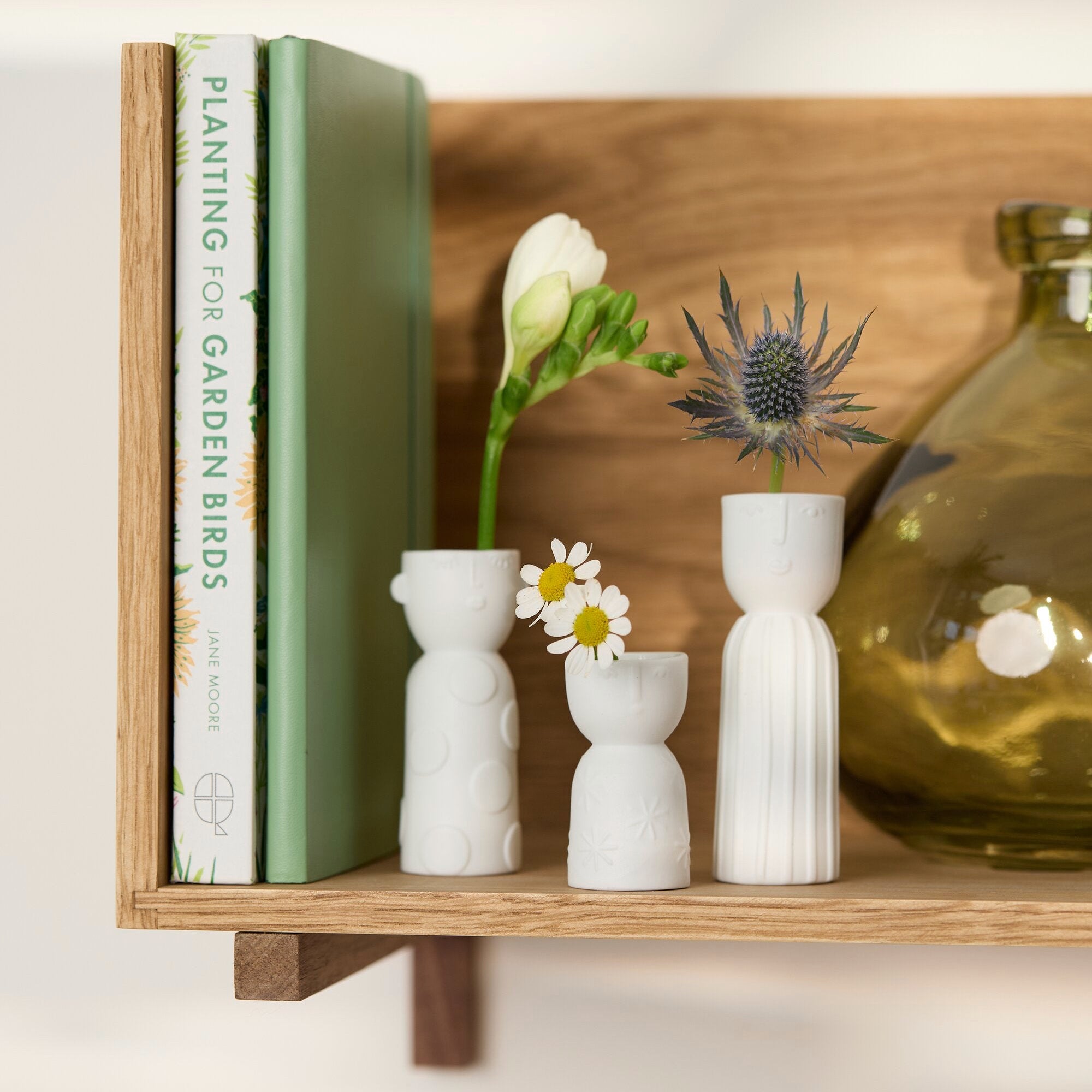 Three small white ceramic bud vases arranged on a wooden shelf, styled with books, flowers, and a glass bottle.