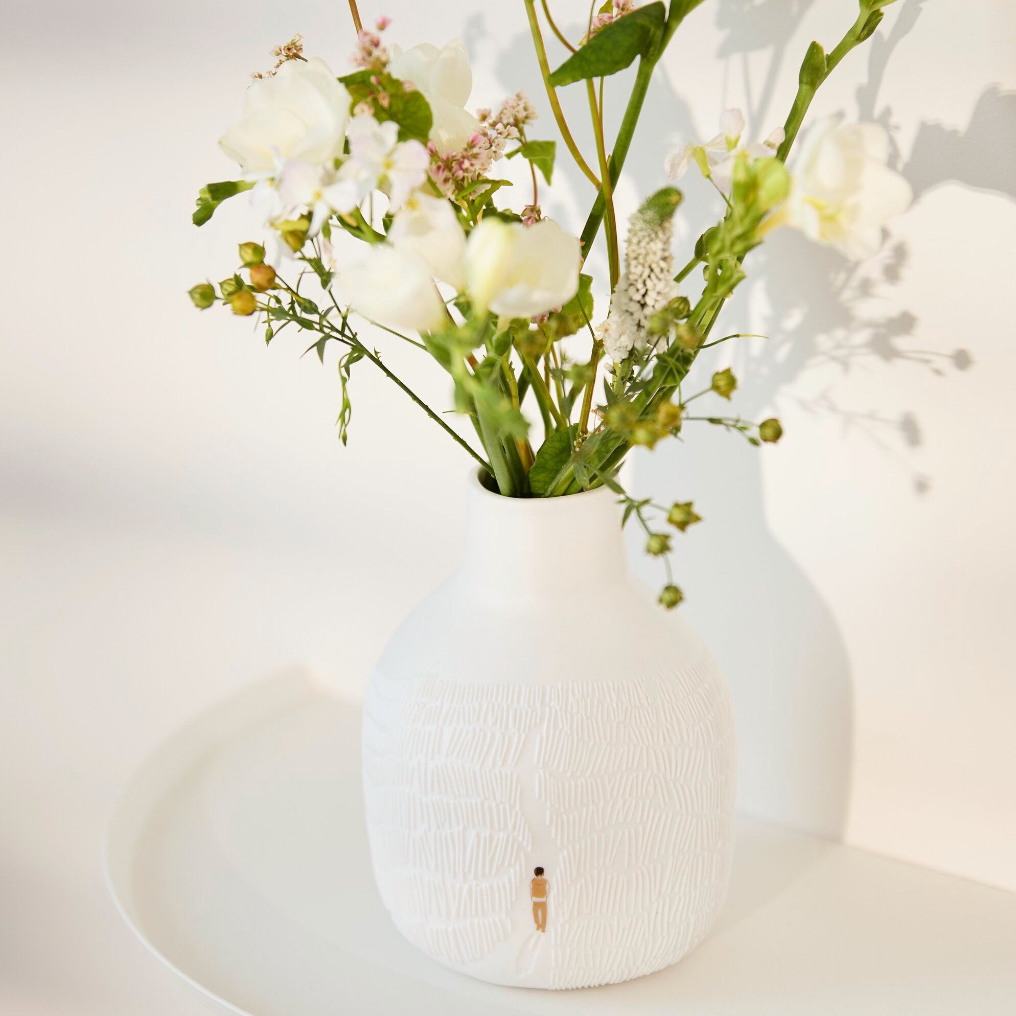 A textured white ceramic vase with a small gold accent, filled with white and pale yellow wildflowers and green stems, set on a white tray against a softly lit white background.