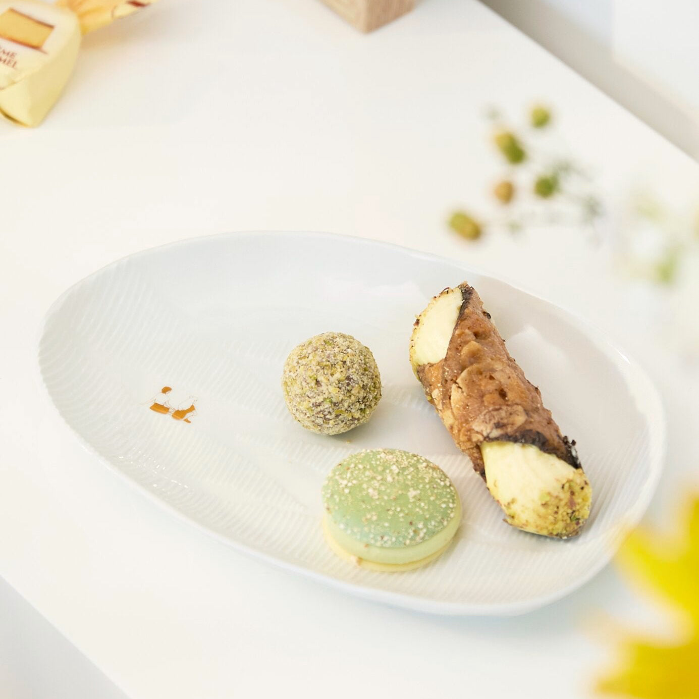 A white oval plate holding three pastries on a bright white tabletop: two green macarons and a sliced pastry with cream and fruit filling, styled with soft blurred background elements.