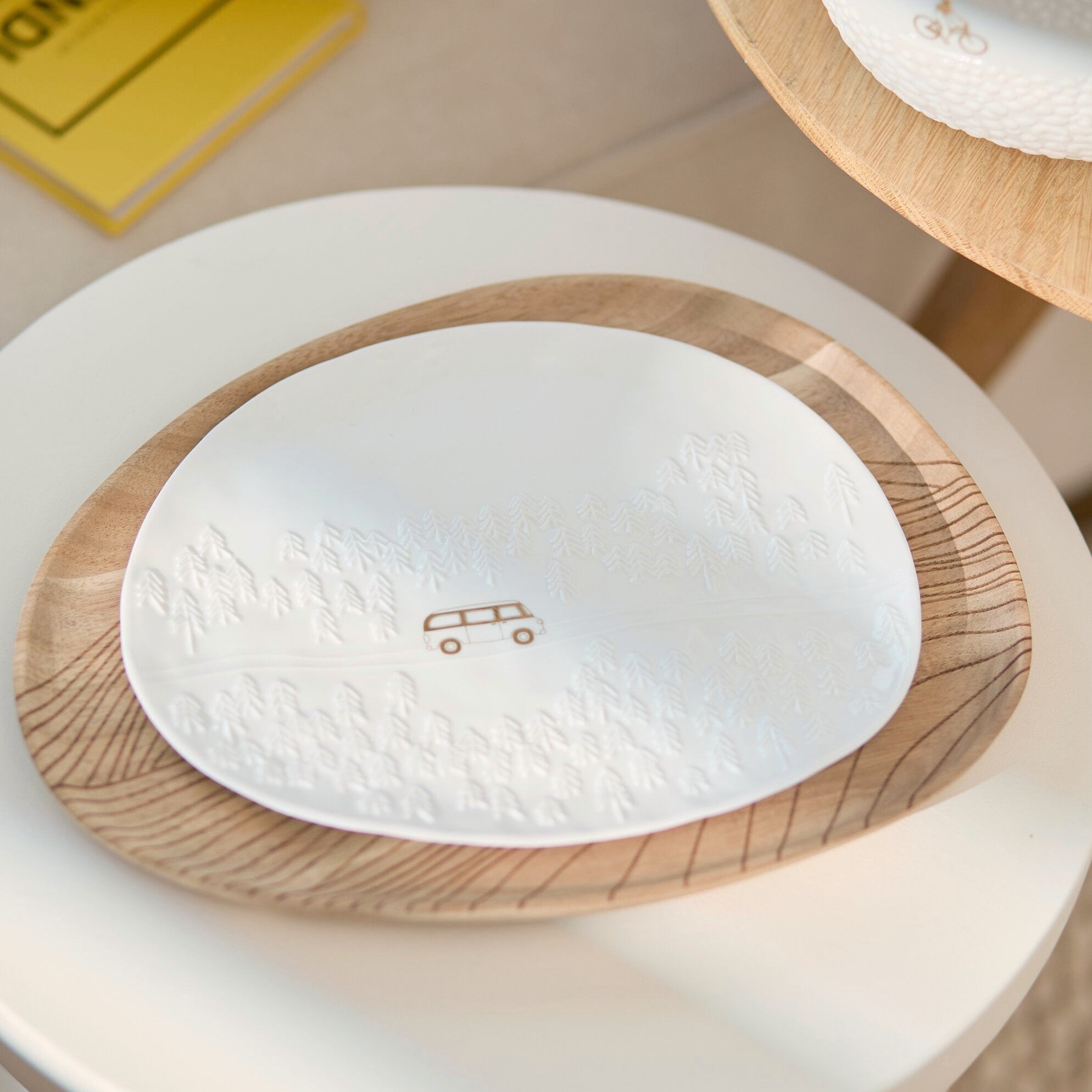A white oval ceramic plate with a textured embossed pattern and a small bus icon in the center, resting on a round wooden tray over a white pedestal.