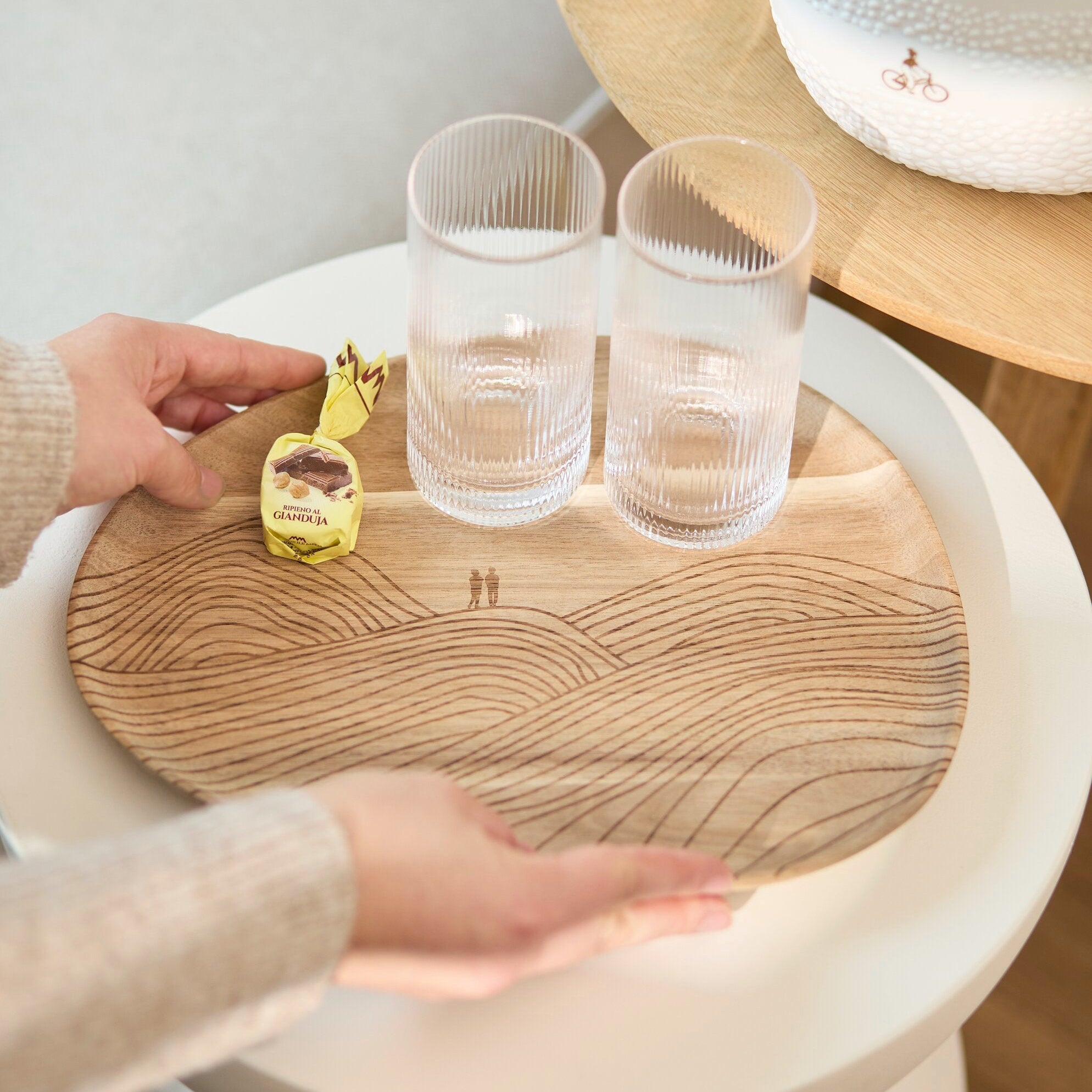 A round wooden serving board with engraved wave-like patterns is placed on a white side table. Two clear textured drinking glasses sit on top, along with a small yellow wrapped item.
