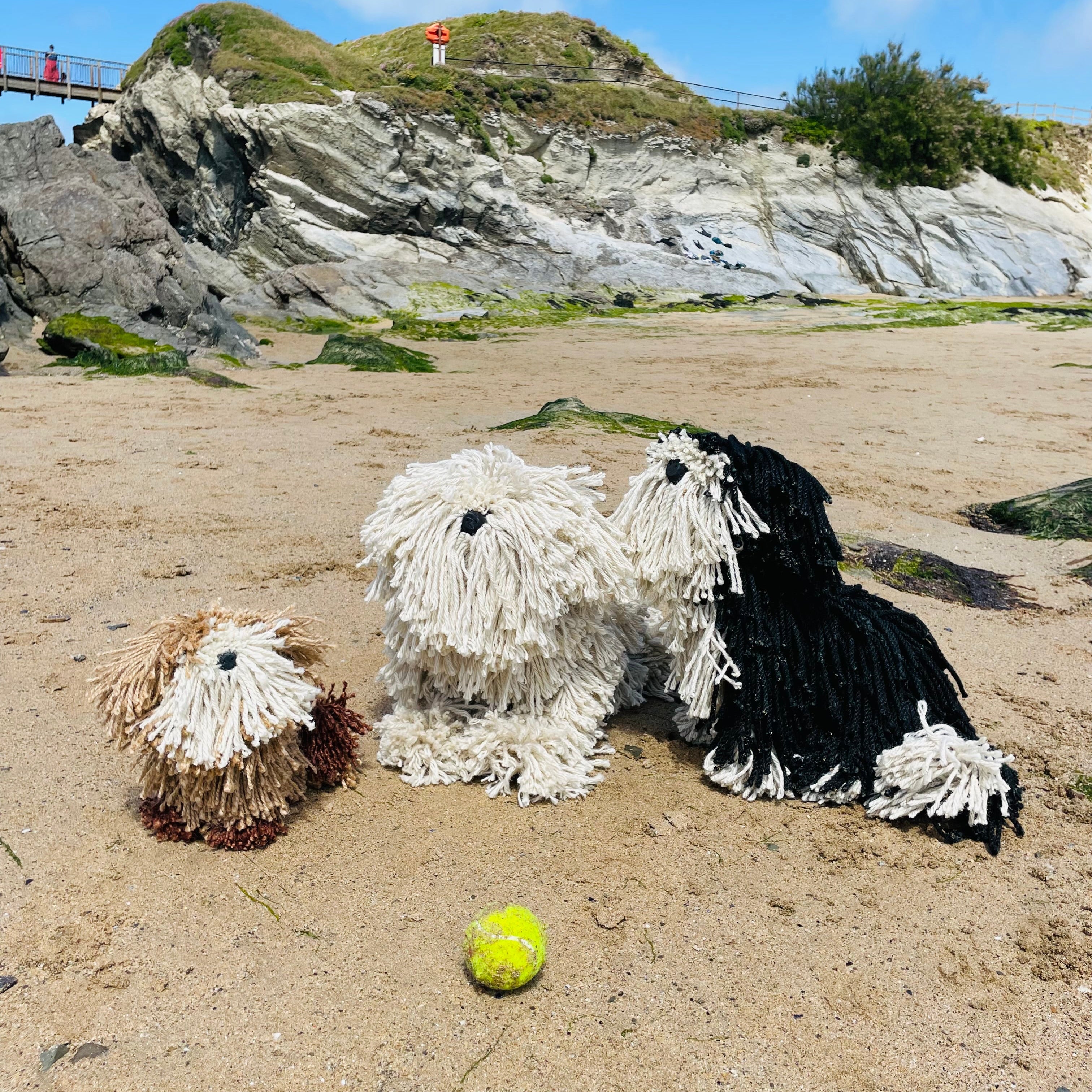 Three fluffy dog sculptures on a beach with a yellow ball and a rocky hill in the background.