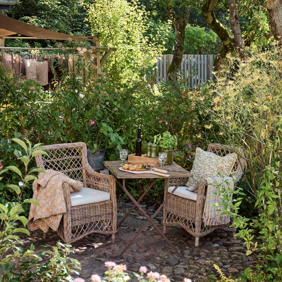 Garden setting with wicker furniture, table, and cushions surrounded by greenery.