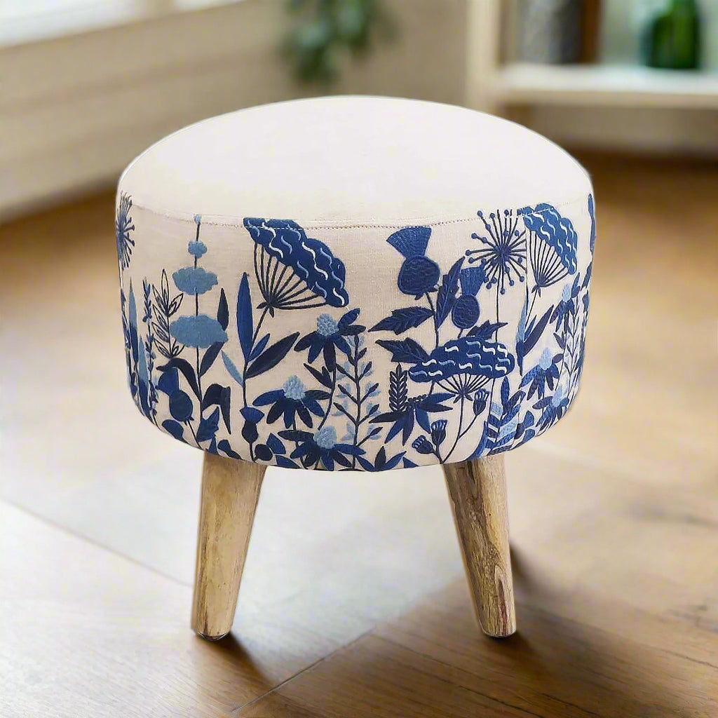 A round footstool with blue floral embroidery on a beige background, featuring a flat base with three wooden legs.