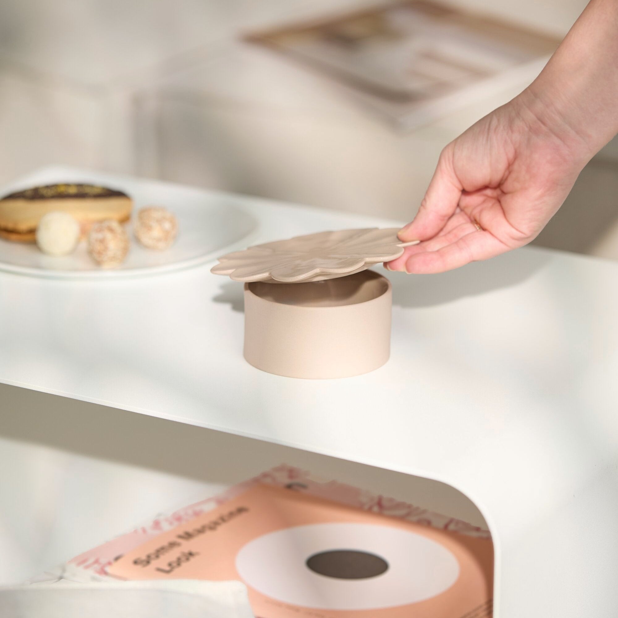 A hand lifts the lid of a small beige round container placed on a white shelf, with a plate of pastries and a stack of books or magazines nearby in a softly lit indoor setting.