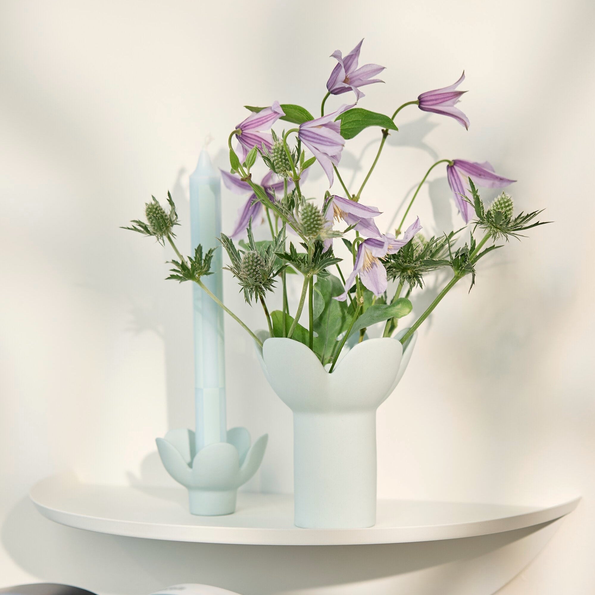 A white ceramic flower vase with a flared rim holding light purple clematis stems and green leaves, styled on a white shelf next to a matching white candle holder with a pale blue candle. An open magazine rests on a light beige upholstered chair in the foreground against a softly lit white wall.