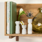 Three small white ceramic bud vases arranged on a wooden shelf, styled with books, flowers, and a glass bottle.