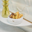 A white textured ceramic bowl filled with cookies and a small chocolate piece, styled on a white shelf next to a translucent yellow glass vase with yellow flowers.