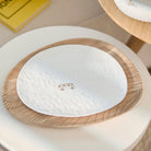 A white oval ceramic plate with a textured embossed pattern and a small bus icon in the center, resting on a round wooden tray over a white pedestal.