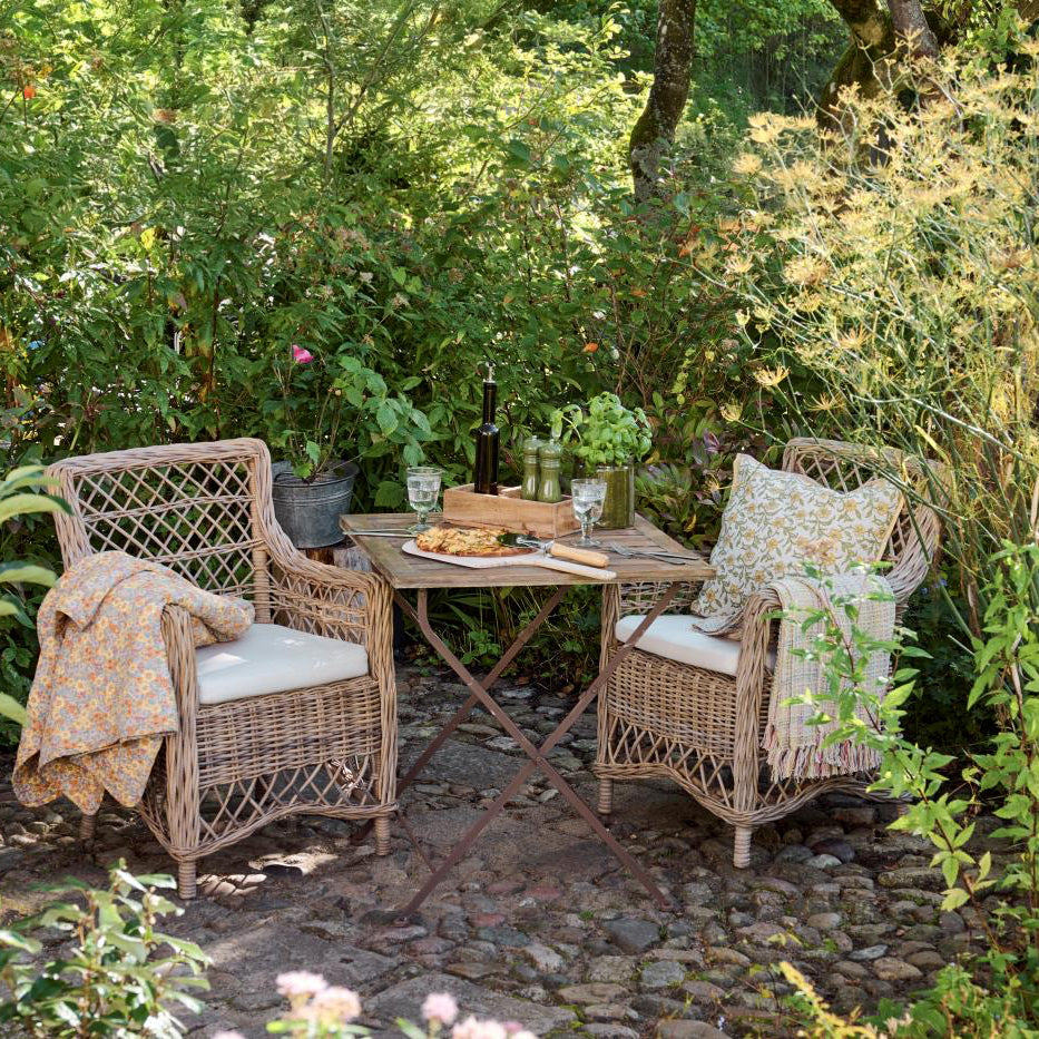 Two wicker chairs with cushions on a stone patio surrounded by greenery