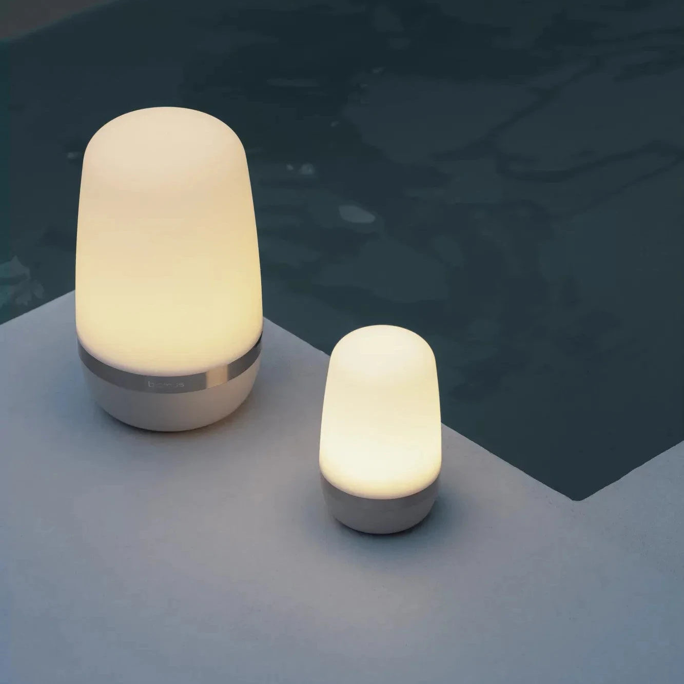 Two illuminated outdoor lamps on a reflective surface with water in the background