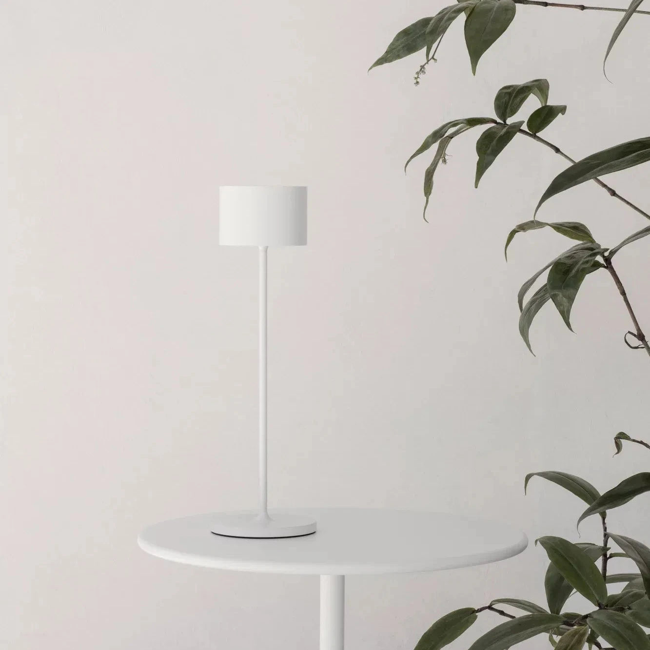 White lamp on a small round table with a plant against a light gray wall