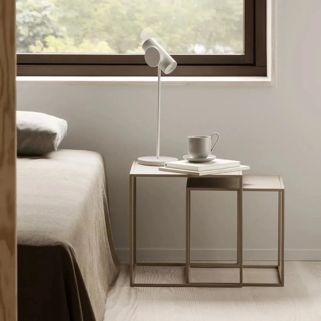 Nest of tables with a cup and lamp near a bed in a room with a window.