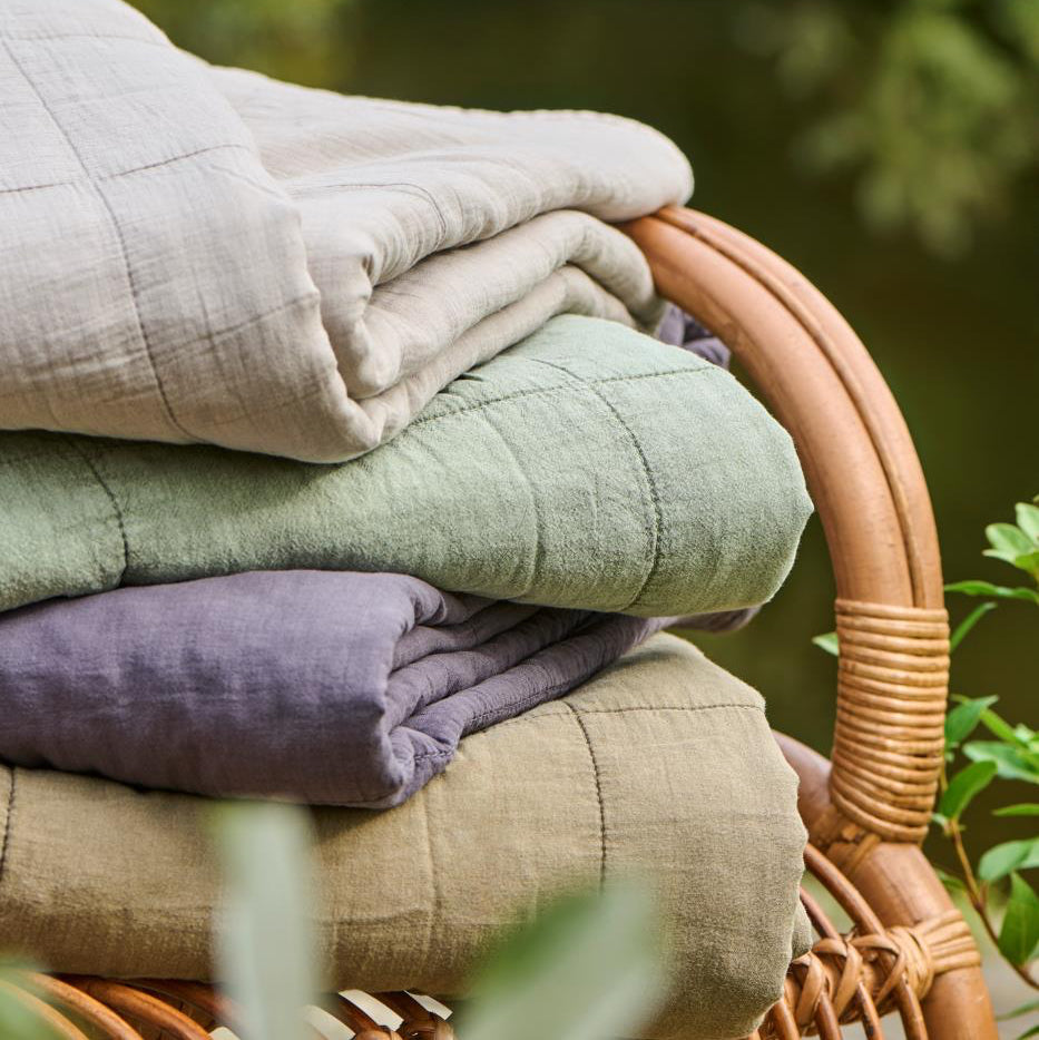Stack of folded fabrics in beige, green, and purple on a wicker chair with a blurred natural background